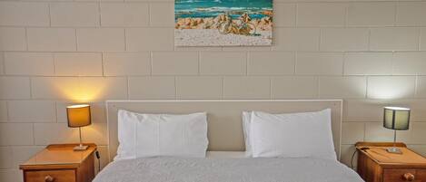 Un dormitorio con una cama, dos mesitas de noche, una lámpara y un cuadro de una playa.