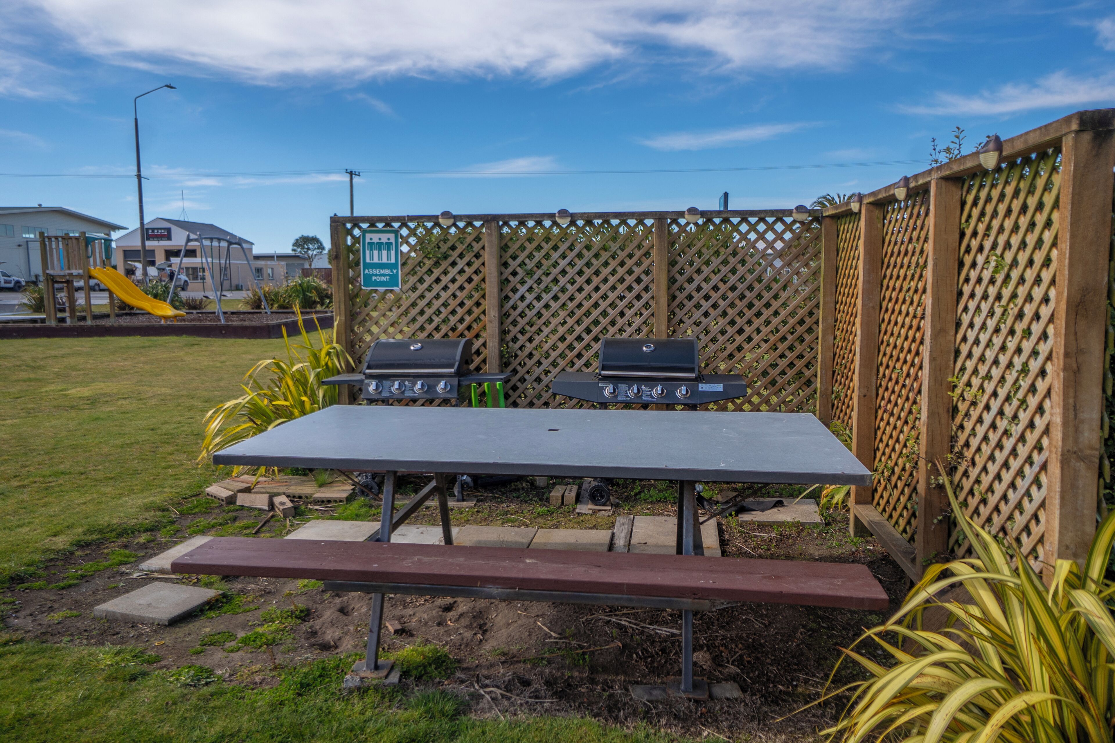 Un área de barbacoa al aire libre con una mesa y dos parrillas, rodeada por una valla de madera y follaje.