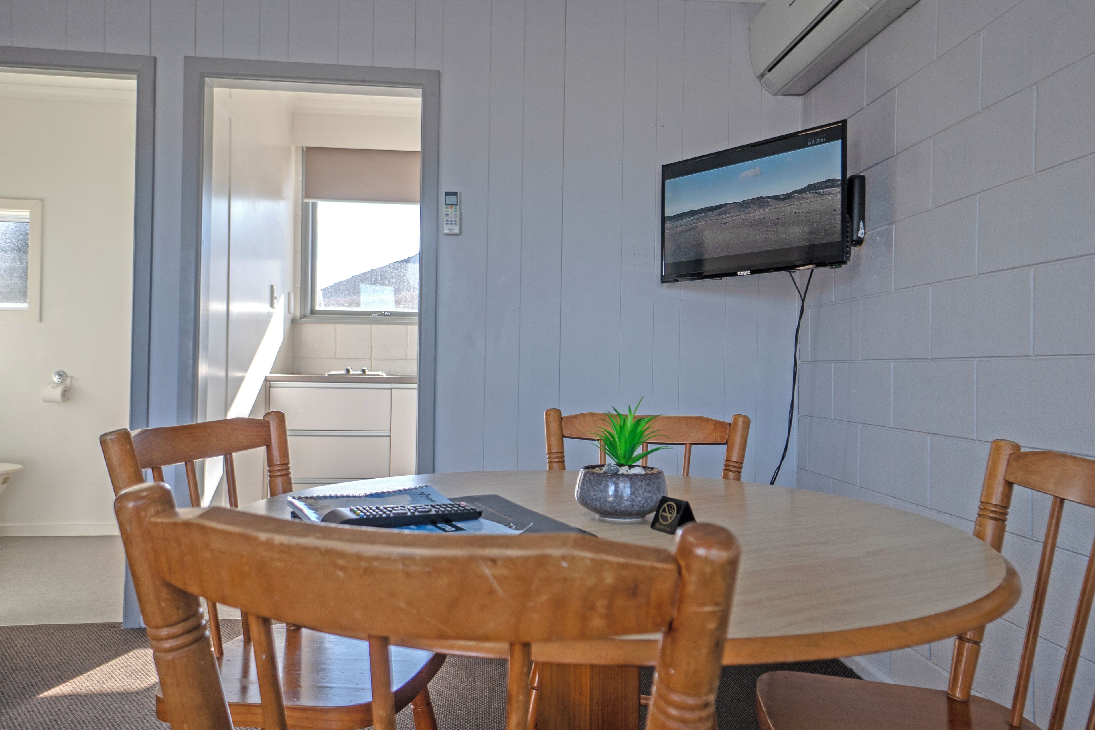 A dining area with a round table, four wooden chairs, a potted plant, a flat-screen TV mounted on the wall, and a door leading to another room.