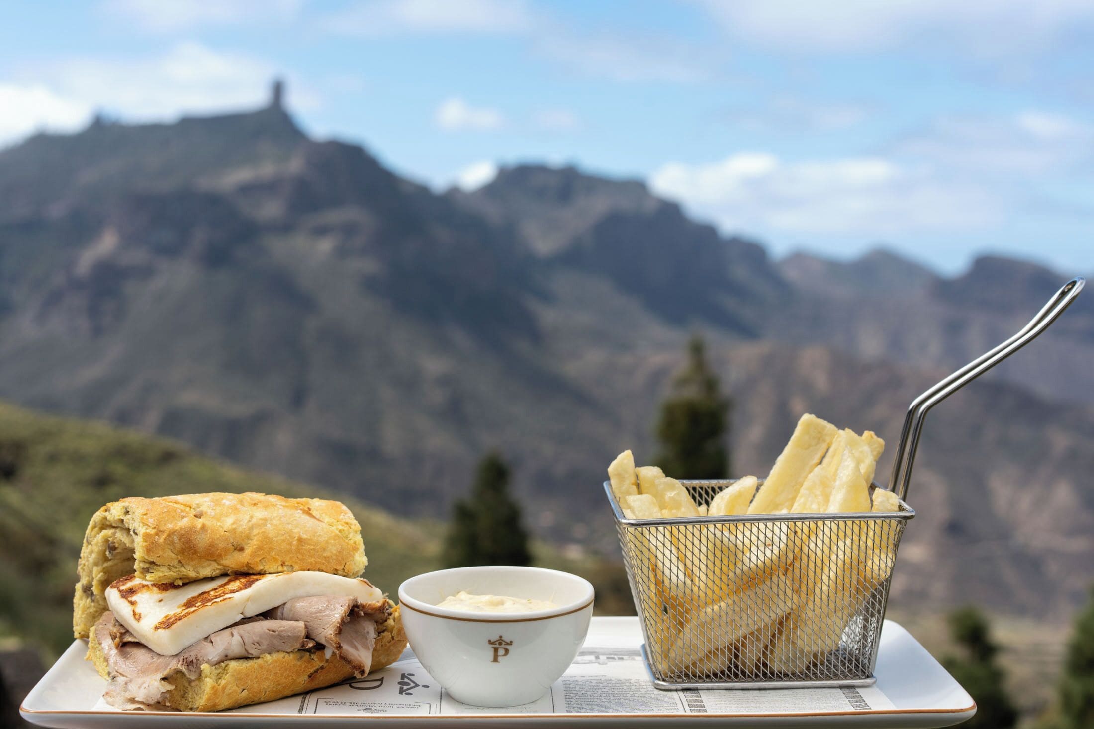 A sandwich, a bowl of dip, and a basket of fries on a table with a mountainous backdrop.