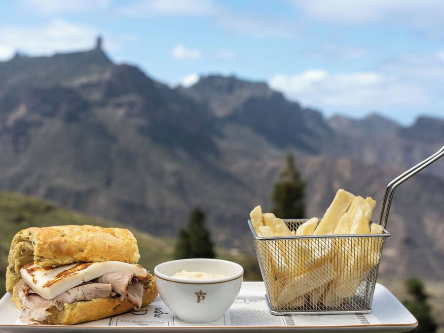A sandwich, a bowl of dip, and a basket of fries on a table with a mountainous backdrop.