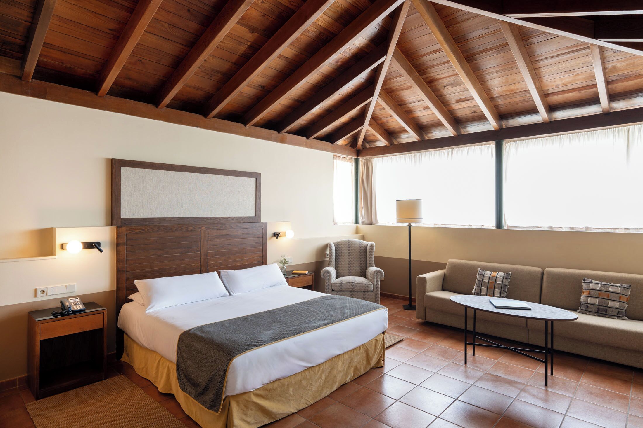 Una habitación de hotel con techo de madera, una cama, un sofá, una mesita y una silla.