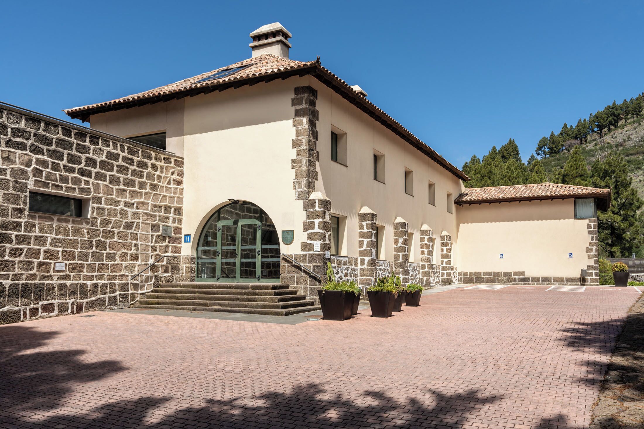 A building with a stone facade, a tiled roof, and a paved courtyard with potted plants.