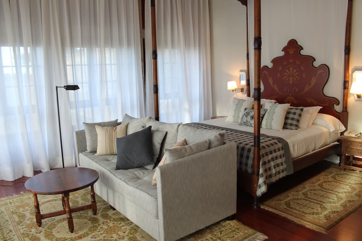 Un dormitorio con una cama con dosel, un sofá, una mesita redonda y un ventanal con cortinas de encaje.