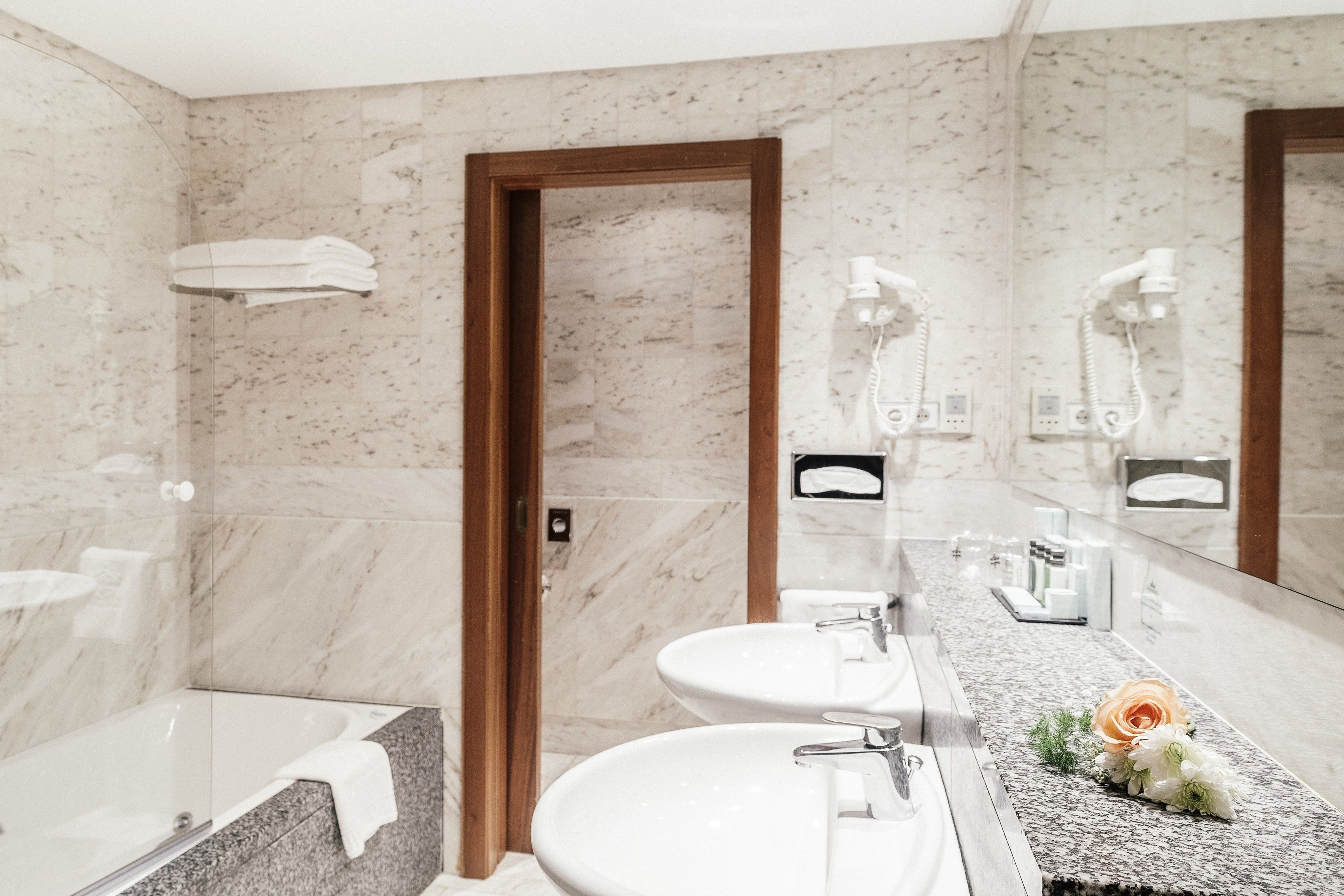 Un baño moderno con pared de mármol, puerta de madera, bañera, dos lavamanos y tocador con espejo.