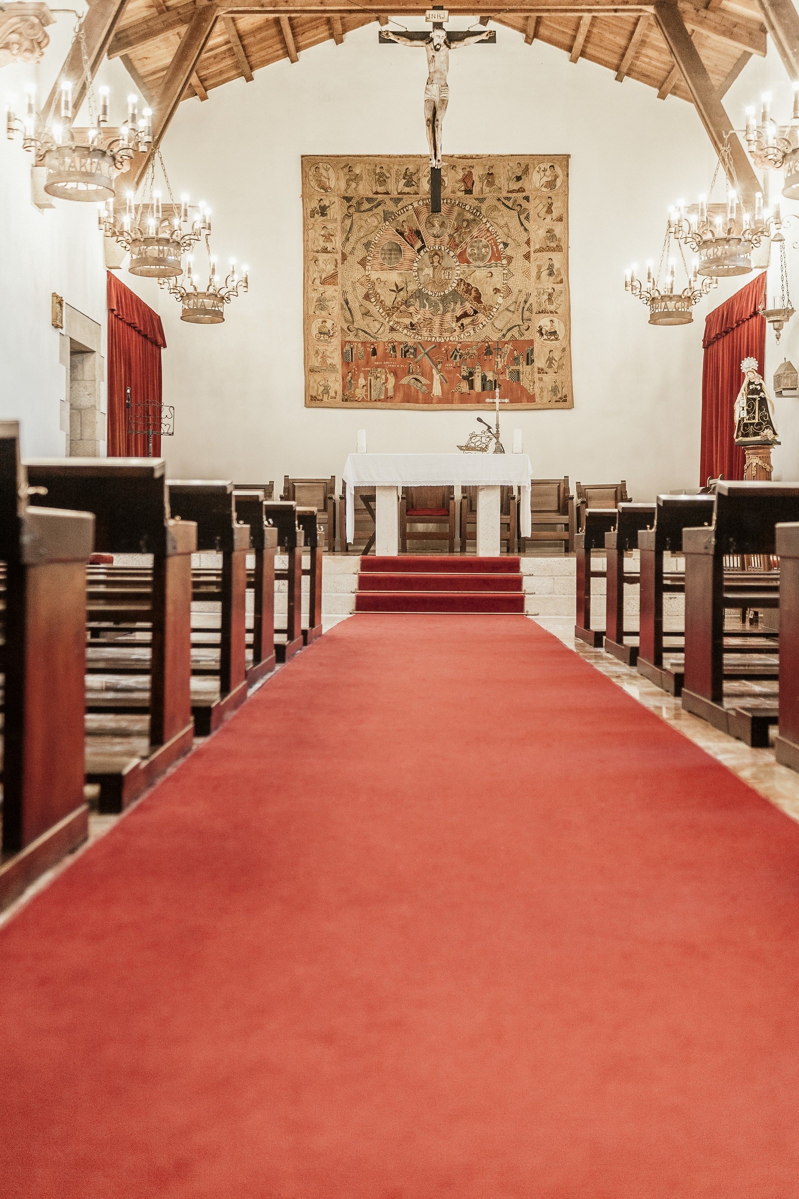 An interior of a church with a red carpet aisle, wooden pews, and a large religious tapestry on the wall.