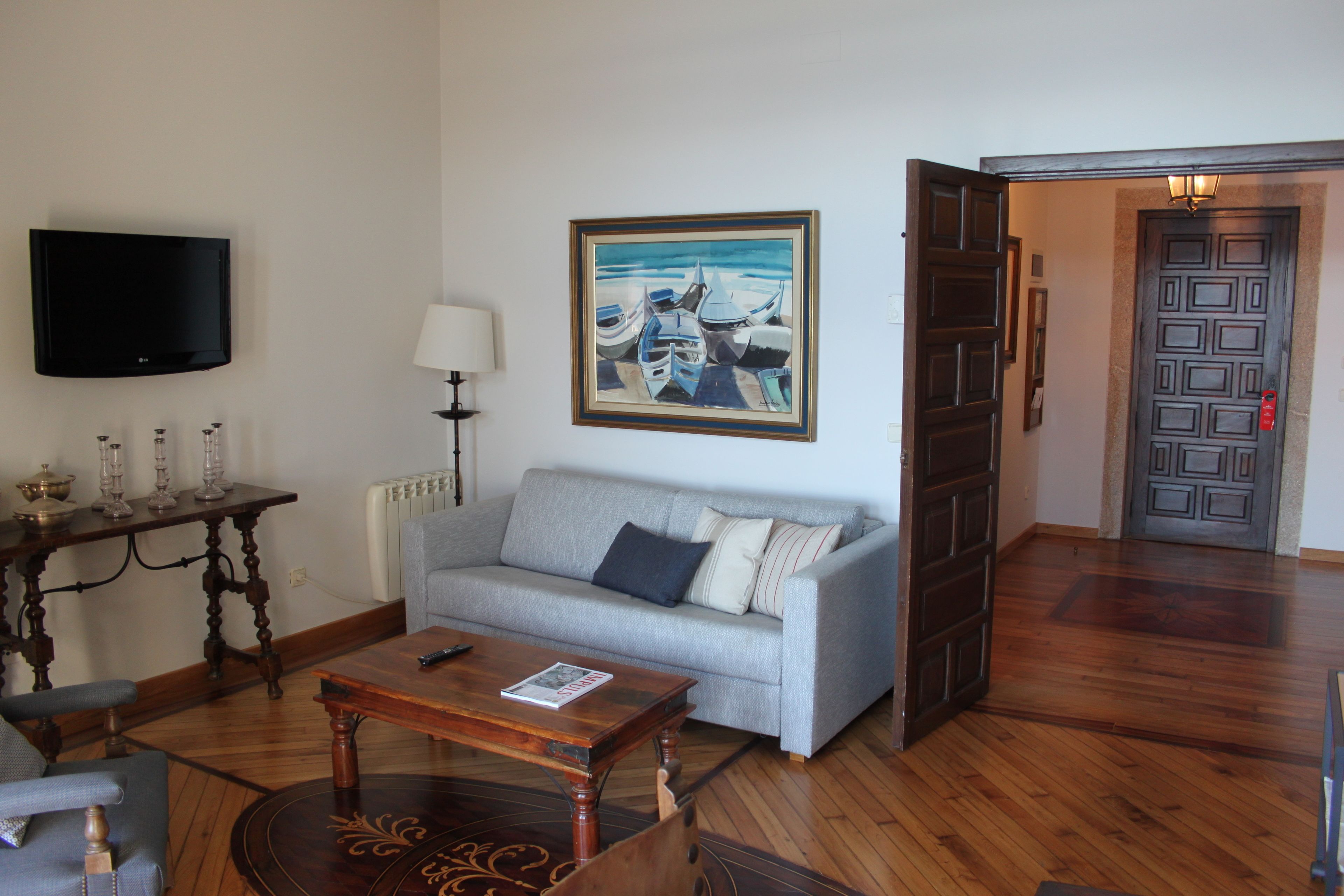 Un salon avec un canapé gris, une table basse en bois, un téléviseur fixé au mur et un tableau représentant des bateaux.