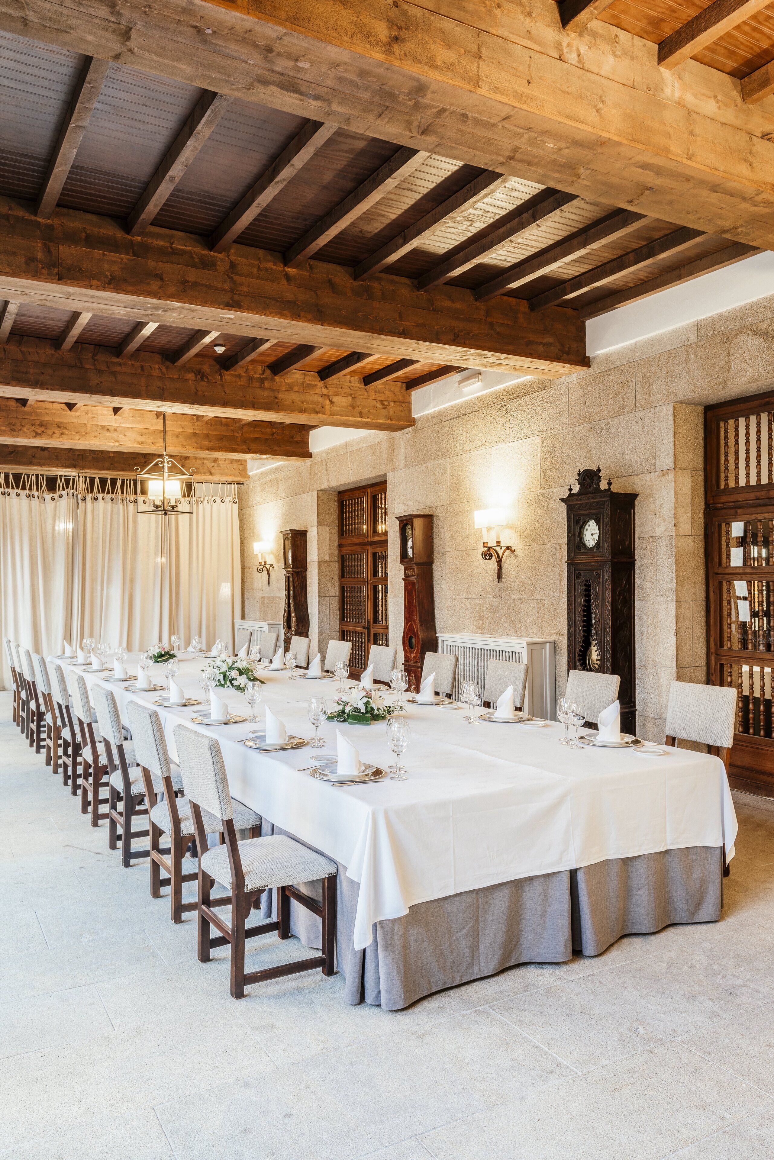 Una larga mesa de comedor preparada para una comida formal, con sillas de madera y un muro de piedra al fondo.