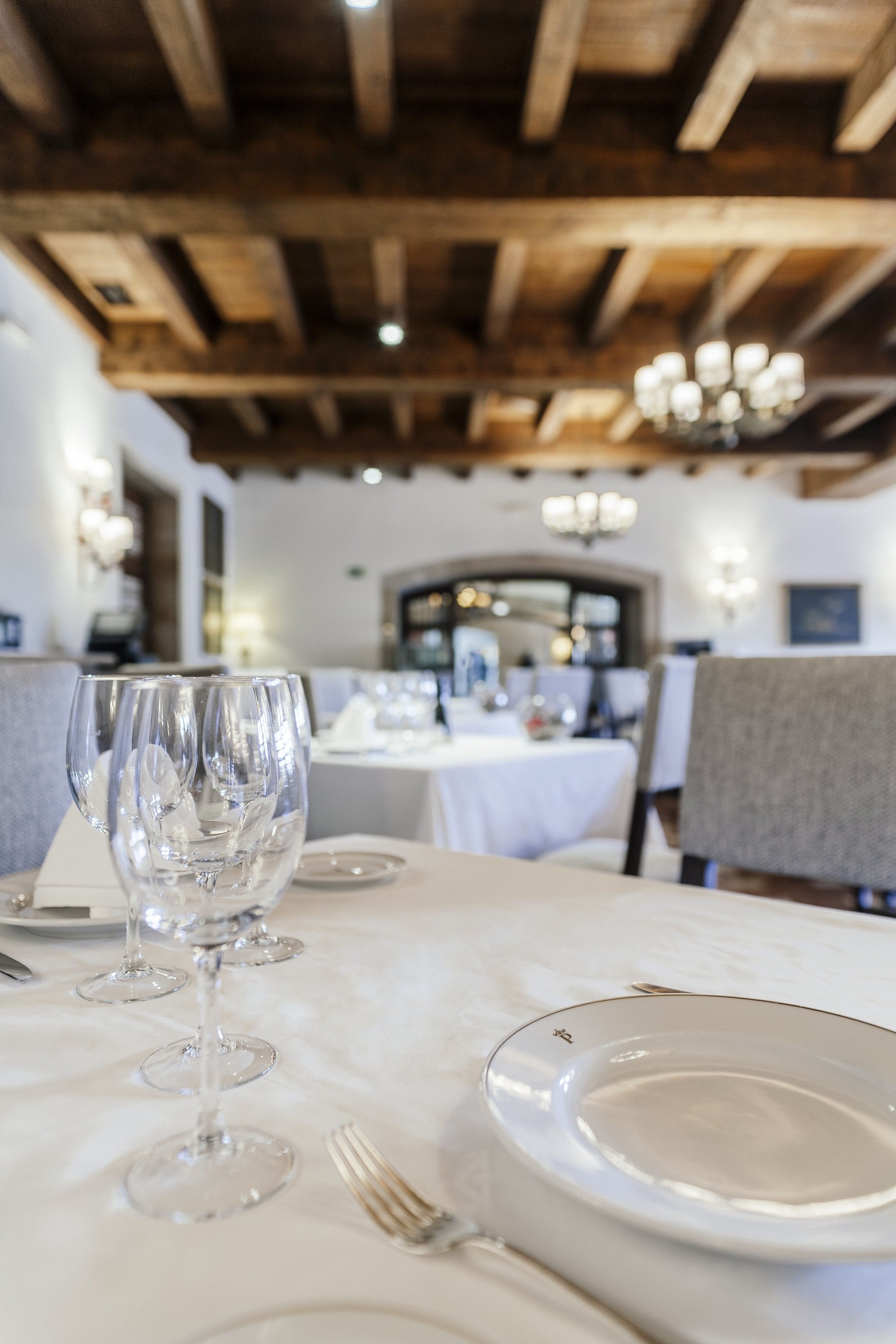 A dining table with a white tablecloth, clear glassware, and a plate with a fork. The room has wooden beams on the ceiling and a chandelier.