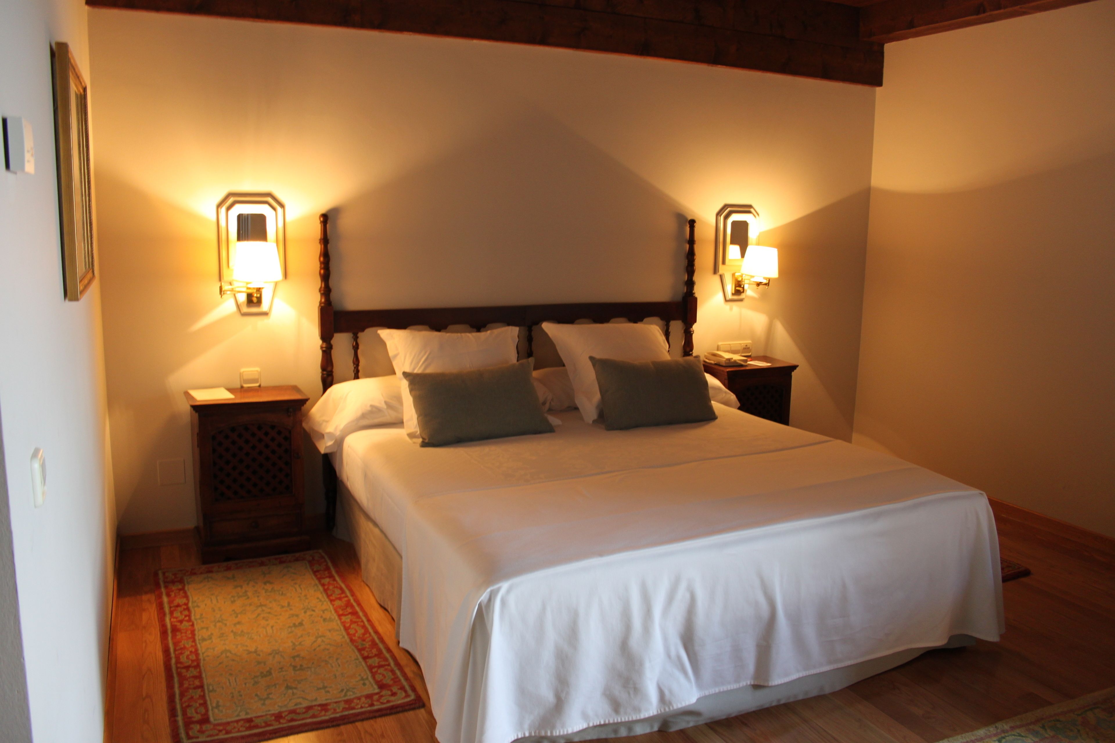 A neatly made bed with white linens and two pillows, flanked by two bedside tables with lamps. A patterned rug is visible on the floor.