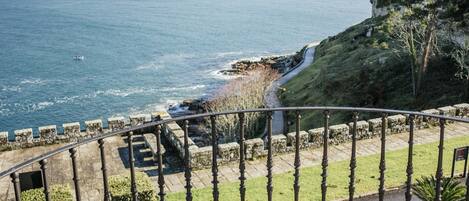 A coastal view from a balcony with a stone railing, a paved path, and a grassy area.