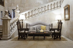 Una imponente escalera con barandillas ornamentadas, un sofá lujoso con cojines, dos sillas de madera y una pequeña mesa con velas.