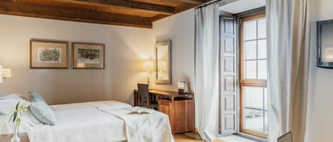 A bedroom with a bed, desk, and laptop. There are two framed pictures on the wall, a lamp, and a window with curtains.