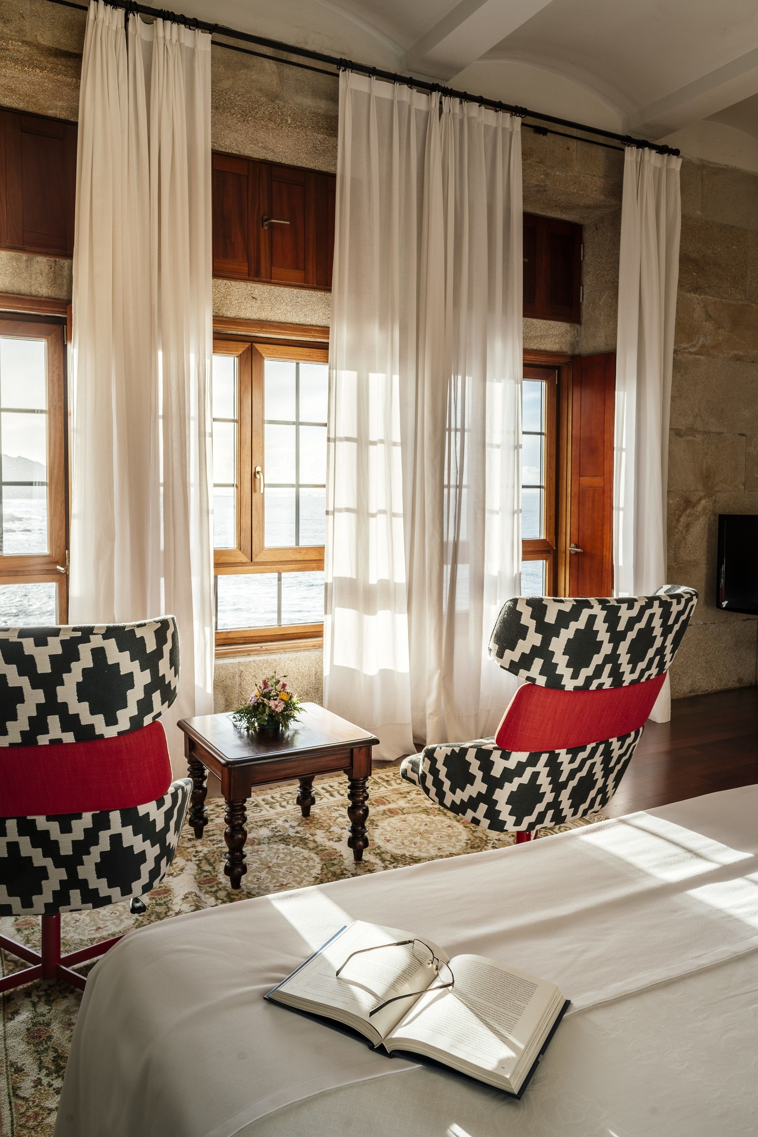 Una habitación con una cama, dos sillones estampados, una mesita con un florero y una ventana con cortinas.