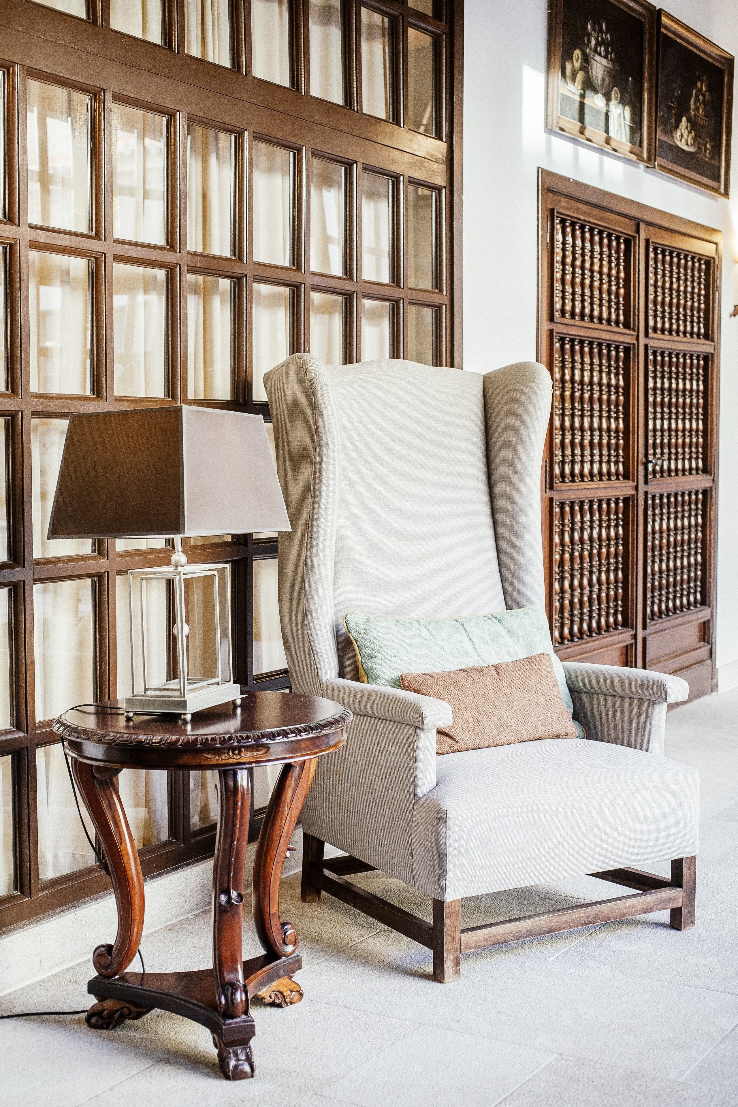 A chair with a cushion, a table with a lamp, and a wooden bookshelf.