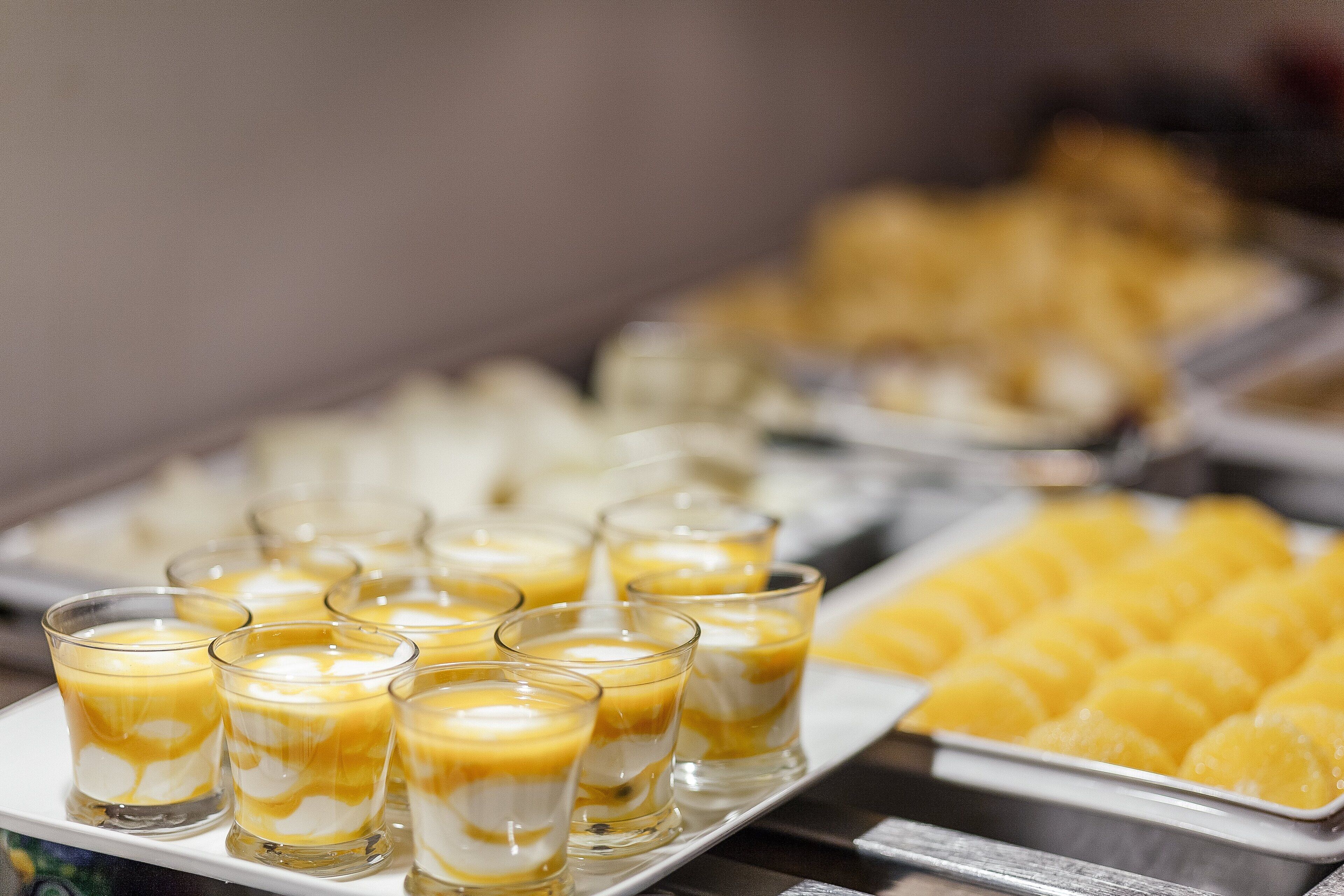 Un bufé con diversos postres, incluyendo una bandeja de cubos de crema pastelera amarilla y varios vasos de postre en capas.
