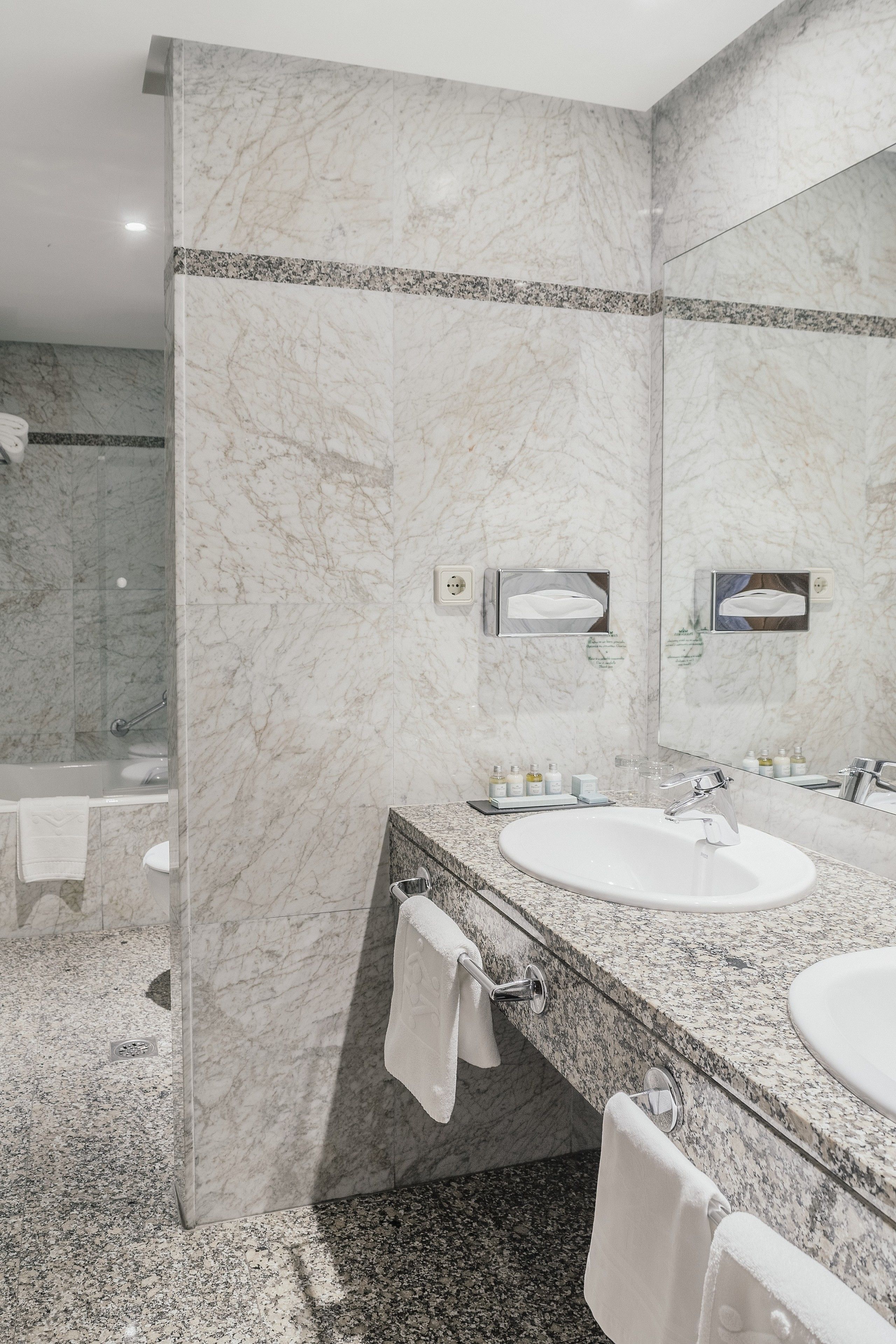 A modern bathroom with marble walls, two sinks, a large mirror, and a glass shower enclosure.