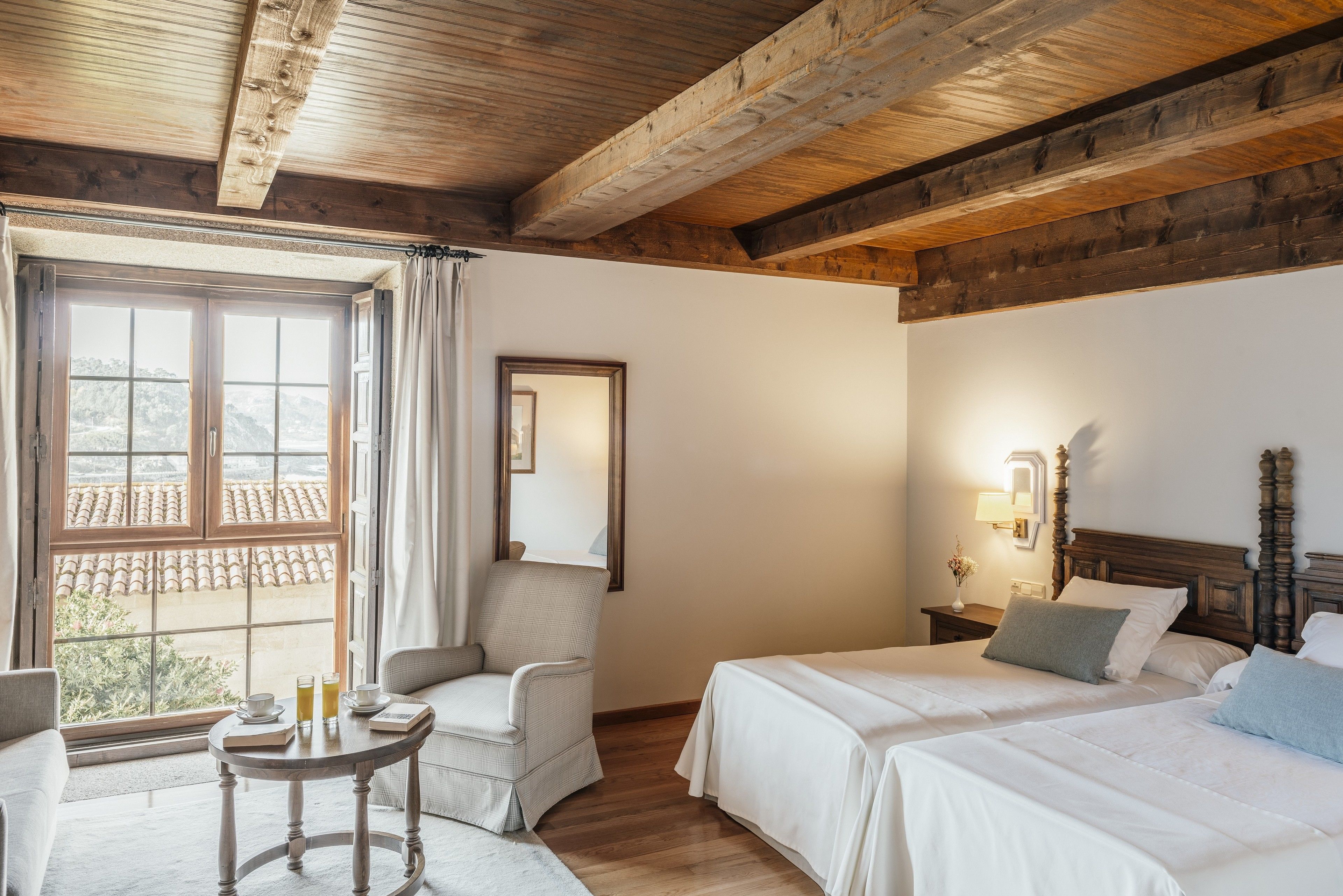 A room with two beds, a chair, a table with drinks, and a window with curtains.