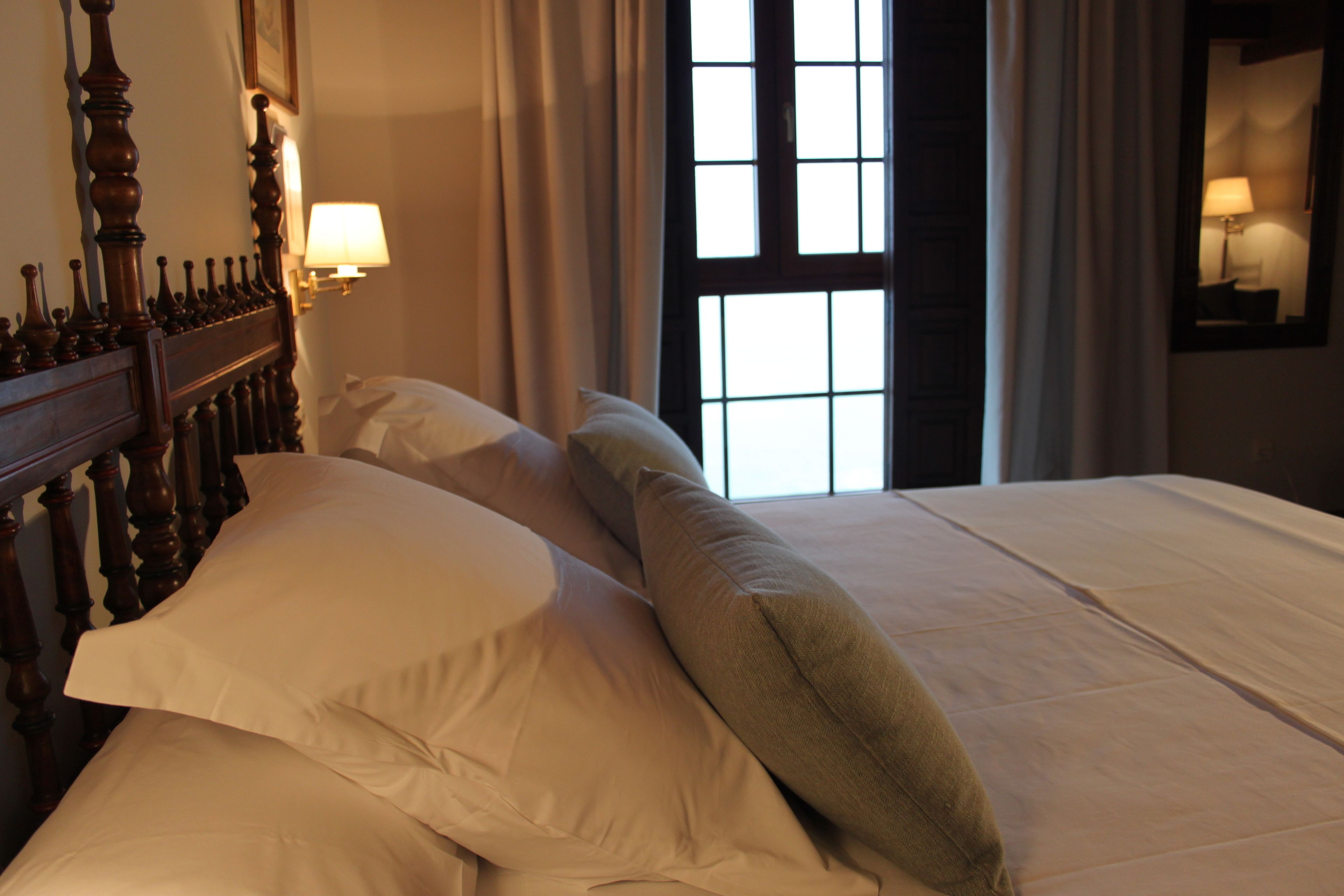 A neatly made bed with a wooden headboard, a window with curtains, and a wall-mounted lamp.