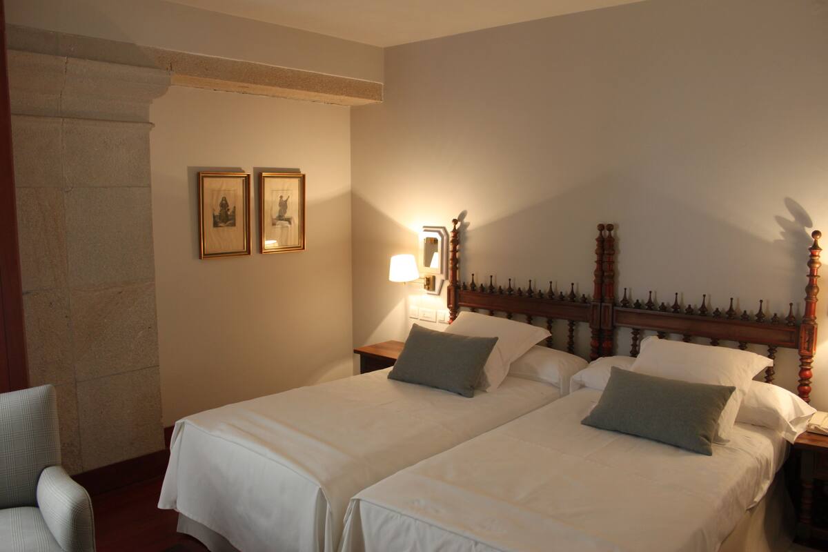 Una cama doble con cabecera de madera, dos mesitas de noche y un tocador. Hay dos cuadros enmarcados en la pared.