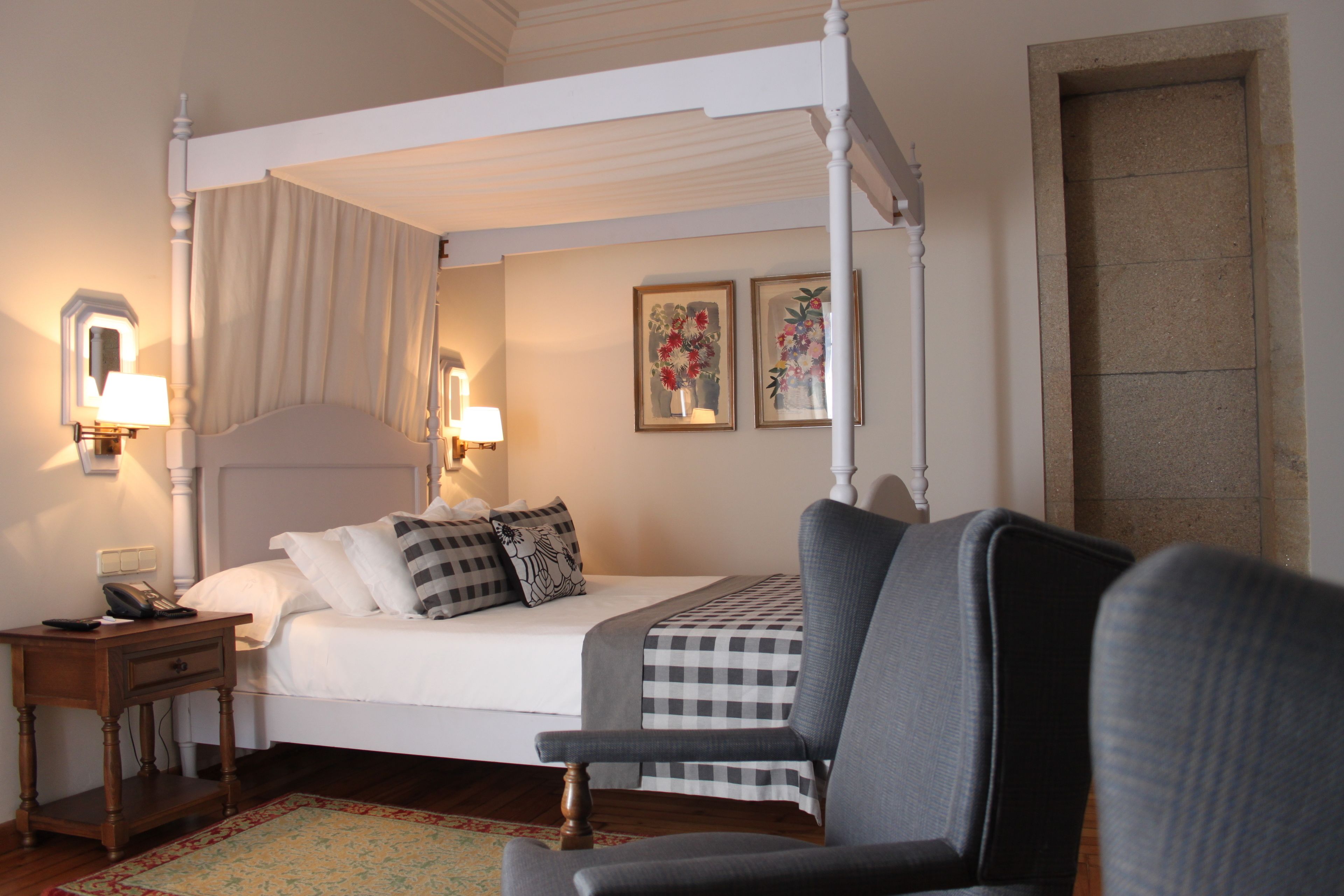 A four-poster bed with a checkered bedspread, a nightstand with a lamp, and two framed floral paintings on the wall.