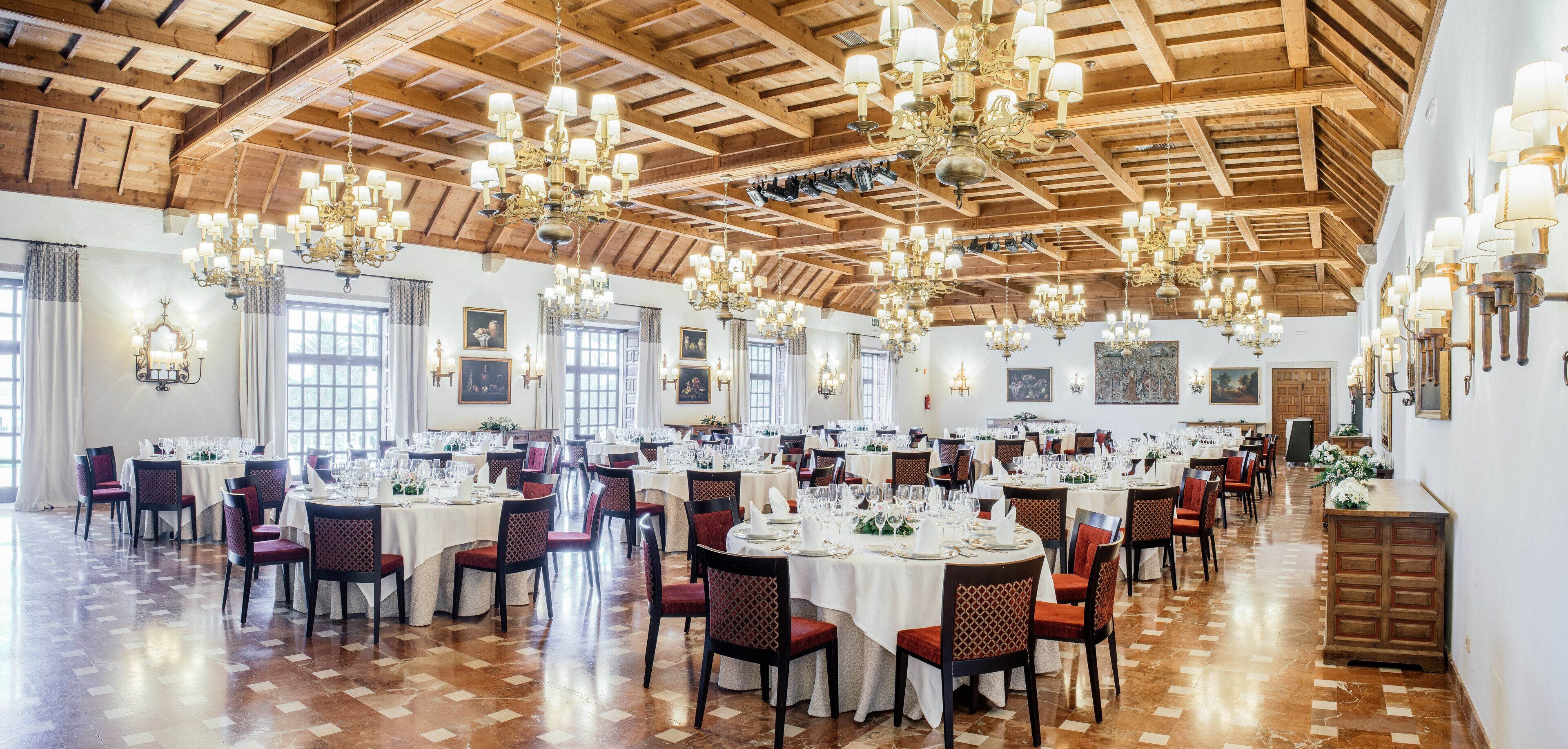 Un amplio comedor con varias mesas redondas dispuestas para un evento formal, vigas de madera en el techo y candelabros.