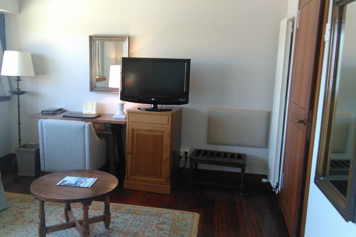 Una habitación pequeña amoblada con un escritorio de madera, un televisor, una silla, una mesita y un radiador.