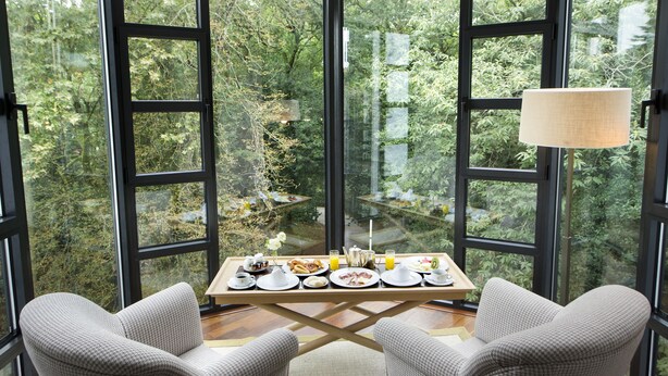 Un comedor con una mesa lista para el desayuno, rodeado de grandes ventanales con vistas a un área boscosa.