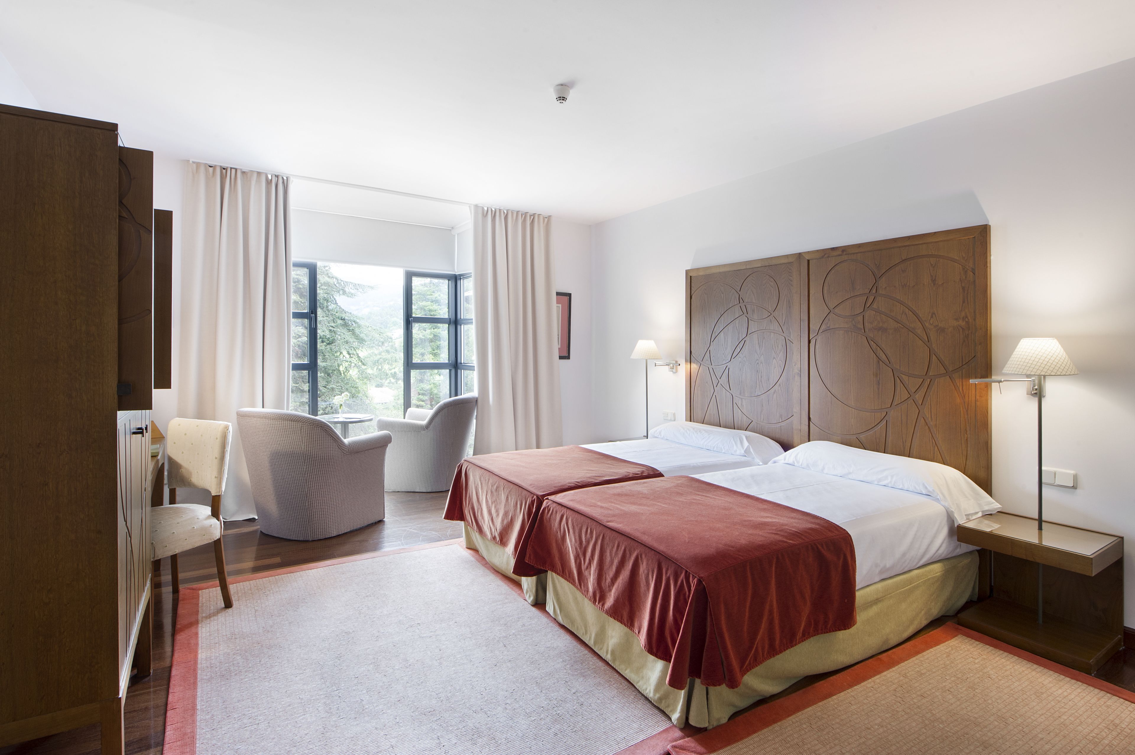 Una habitació d'hotel amb un llit gran, dues làmpades de nit, una cadira, un escriptori i una finestra amb cortines.