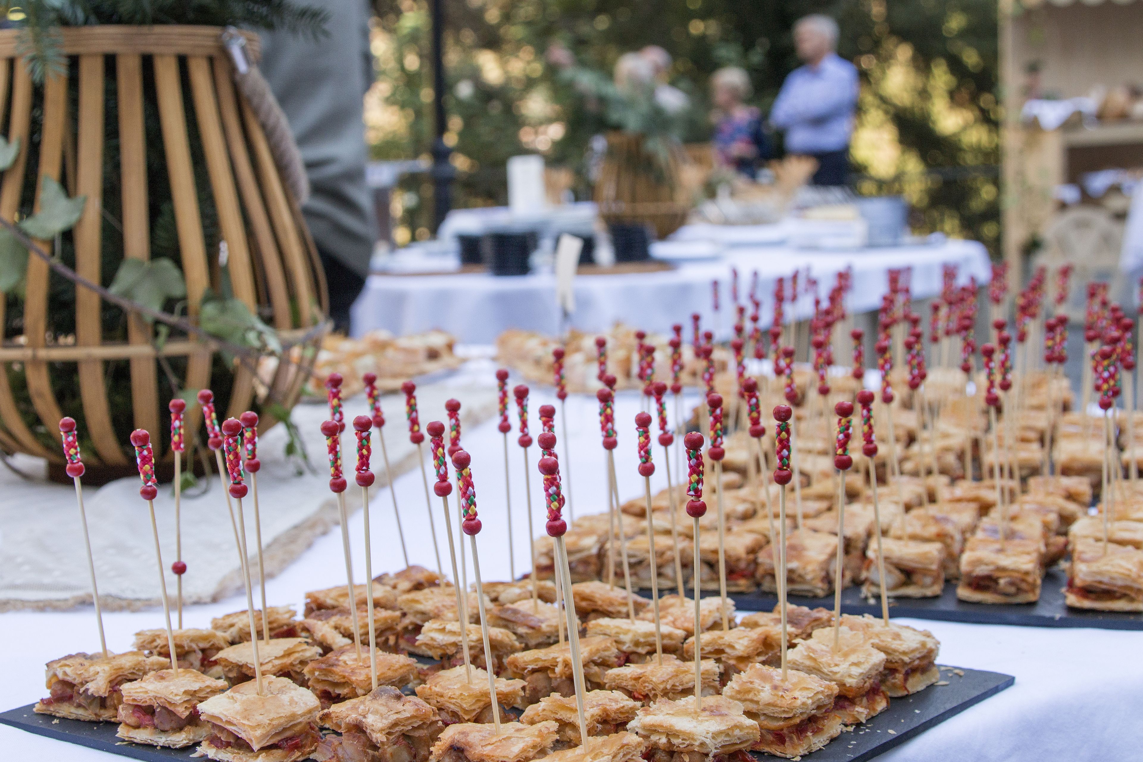A table with square pastries on sticks, a wicker chair, and a blurred background with people.