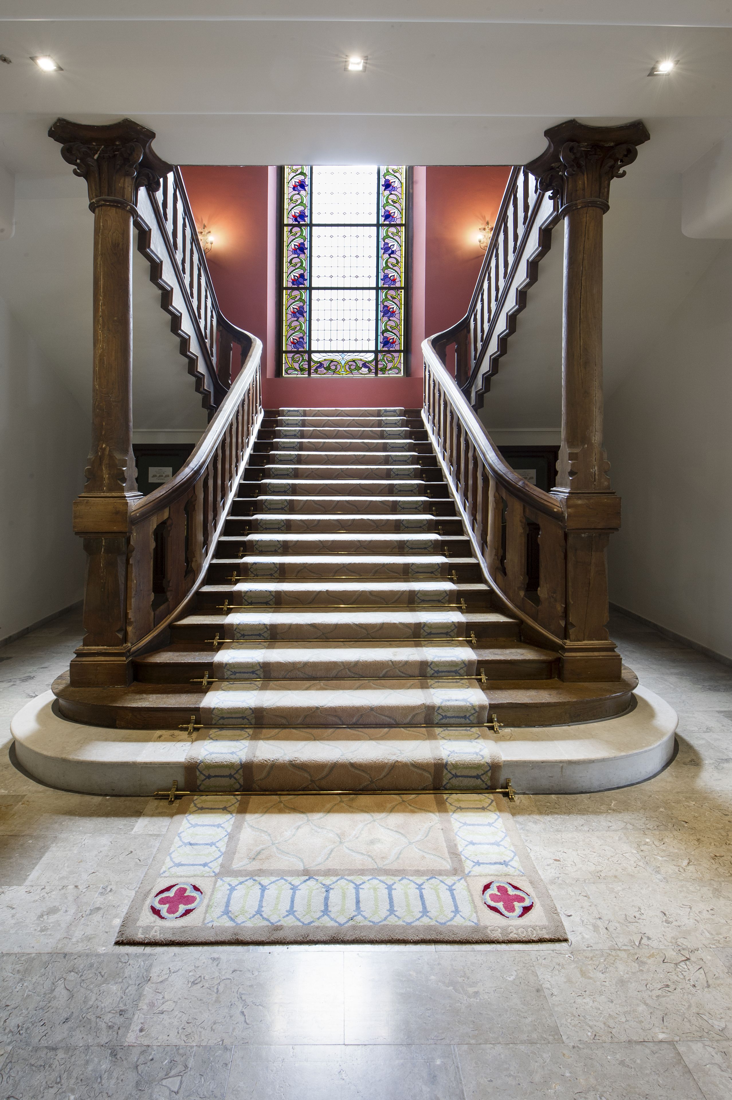 Una gran escalera con barandillas de madera ornamentadas y un suelo estampado que conduce a un vitral.