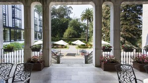 Un espacio patio al aire libre con suelo de piedra, sillas de metal y sombrillas. Hay plantas en macetas y se ve un jardín con árboles y una palmera.