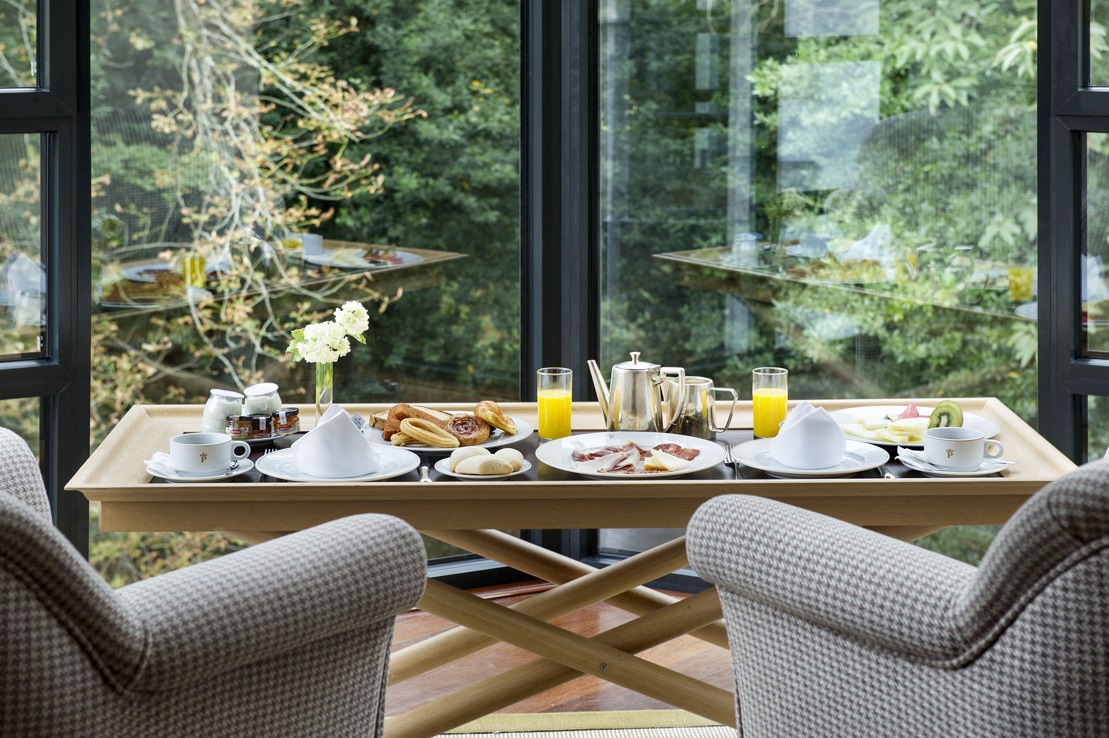 Bandeja de desayuno con diversos platos y bebidas sobre una mesa junto a una ventana con vistas a follaje.