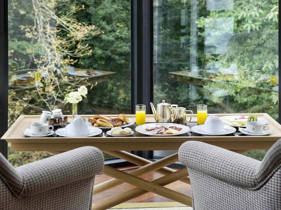 A breakfast tray with various dishes and drinks on a table by a window overlooking greenery.