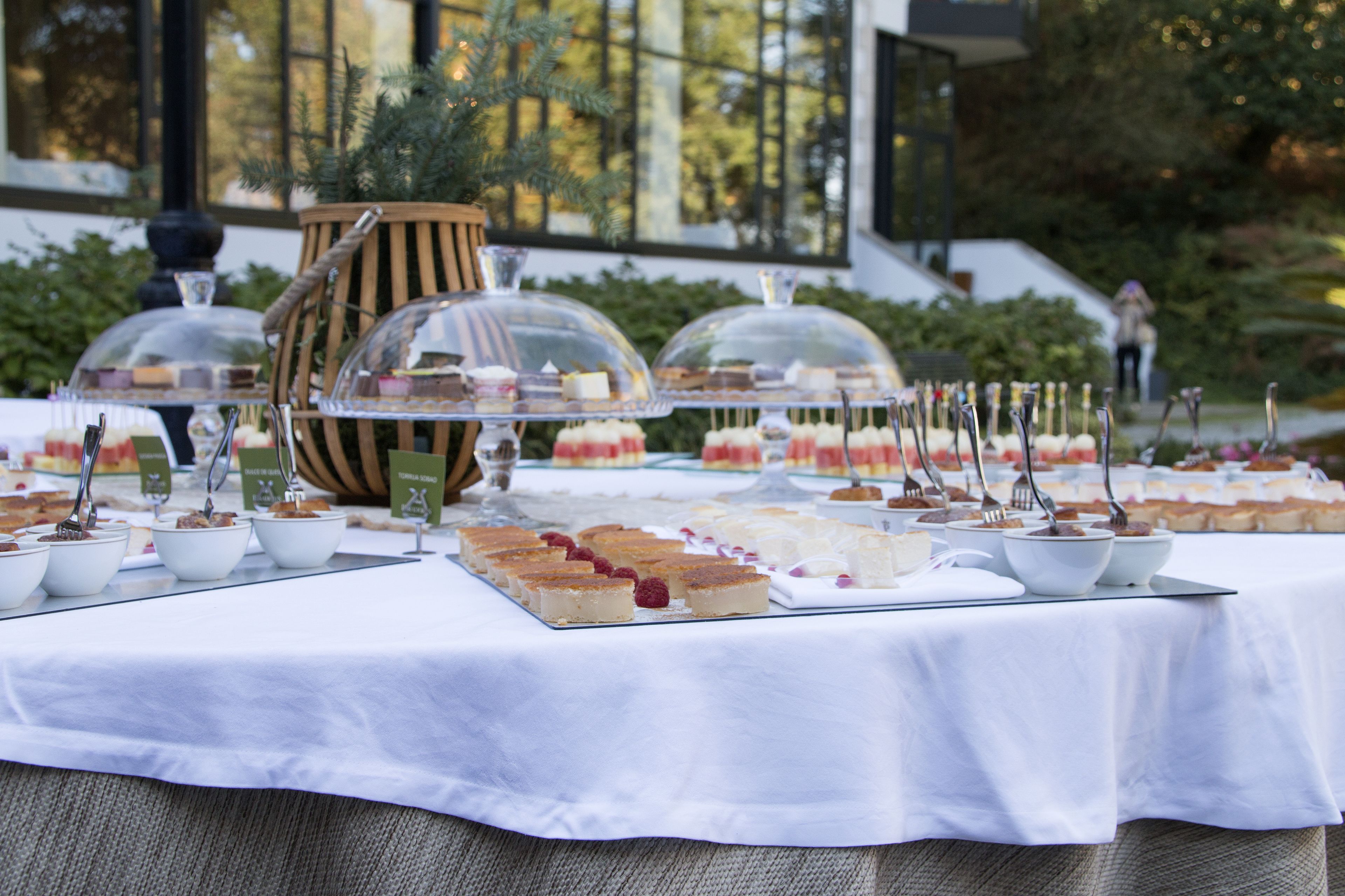 A buffet table with various desserts and pastries, covered with glass domes, set outdoors with a view of a building and greenery in the background.