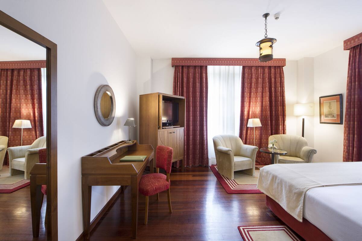 A hotel room with a bed, two armchairs, a desk, a mirror, a wardrobe, and a lamp.