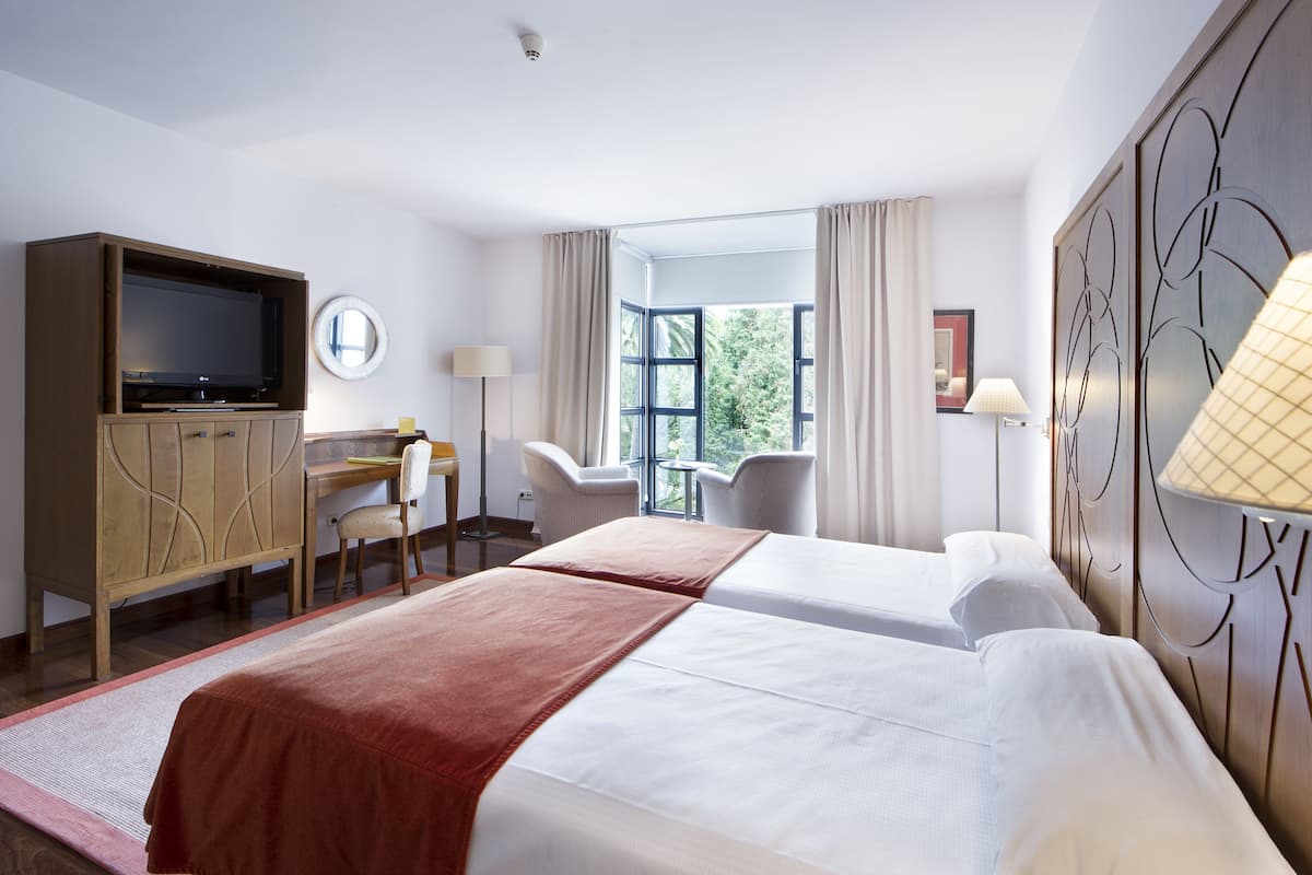 A hotel room with two beds, a desk, a television, and a window with curtains.
