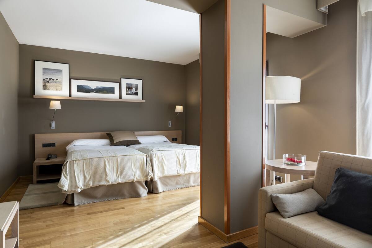 A hotel room with a double bed, bedside tables, a lamp, a small table with a vase, and a sofa.