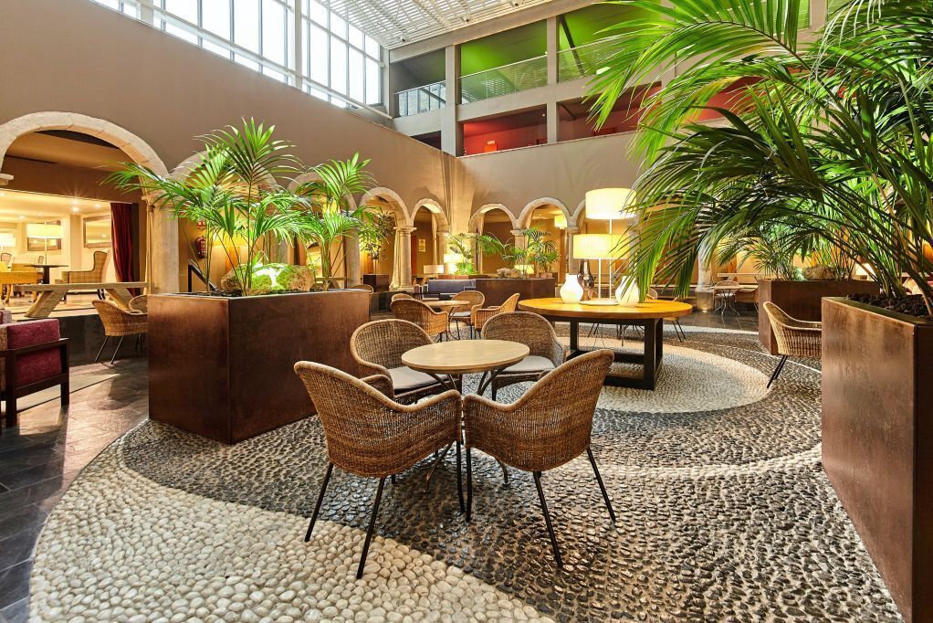 A spacious lobby with a high ceiling, featuring a central seating area with wicker chairs and a round table, surrounded by potted plants and a pebbled floor.