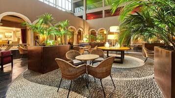 A spacious lobby with a high ceiling, featuring a central seating area with wicker chairs and a round table, surrounded by potted plants and a pebbled floor.