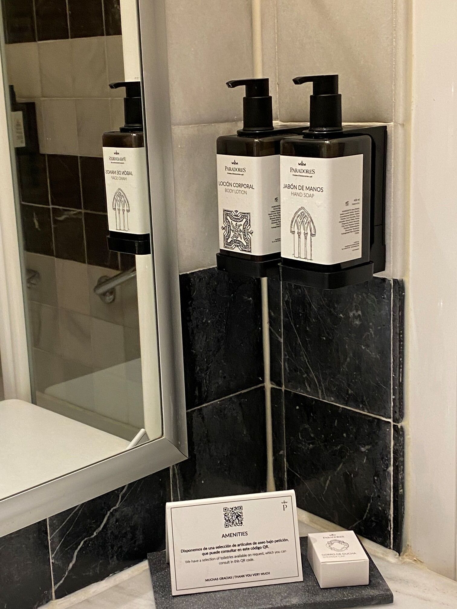 A bathroom with a black marble sink, two bottles of body lotion, and a hand soap dispenser. There is a QR code for amenities and a sign for toiletries.
