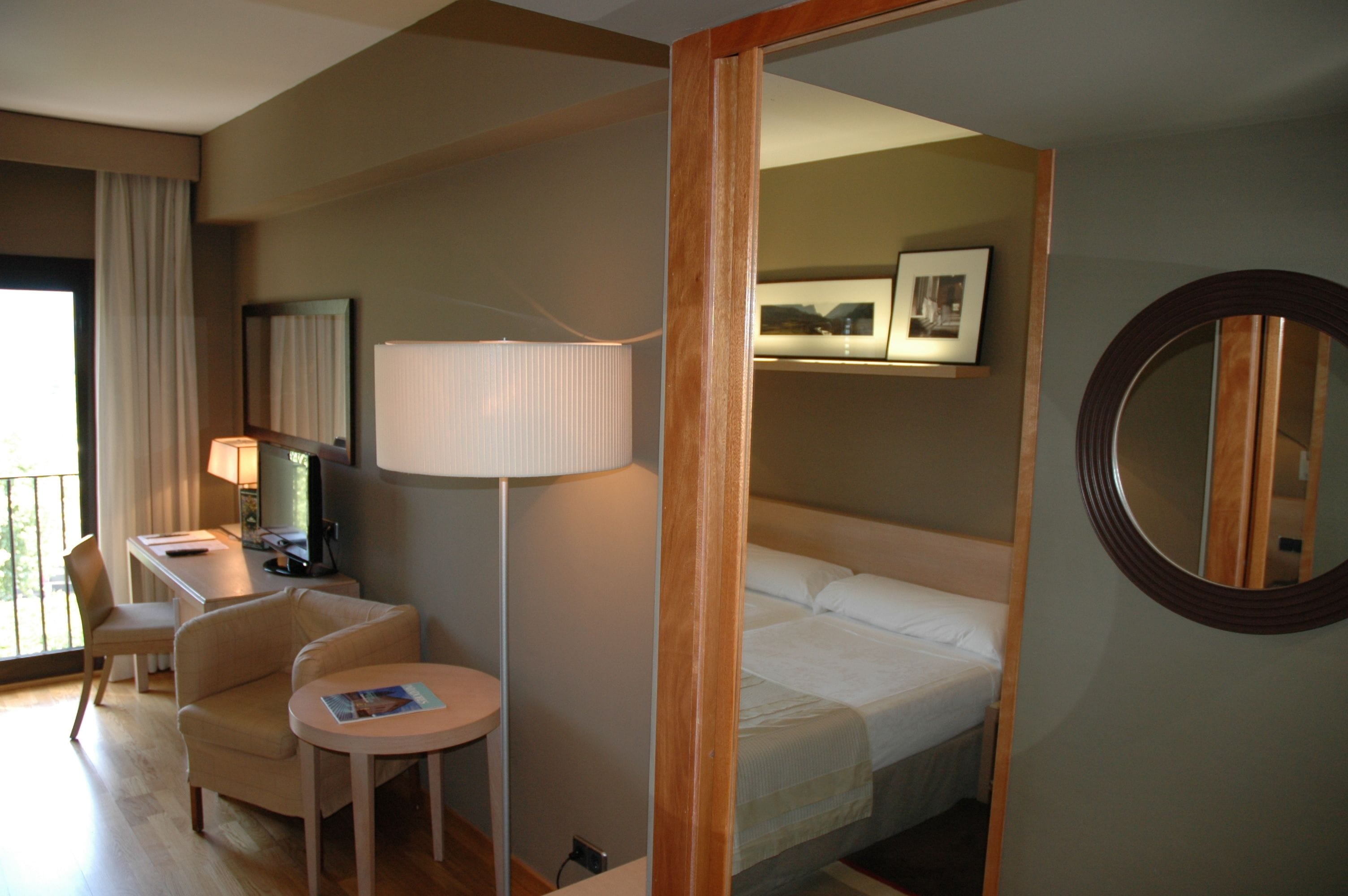 Habitación de hotel con cama, escritorio con computadora, silla, mesita, lámpara y espejo redondo.