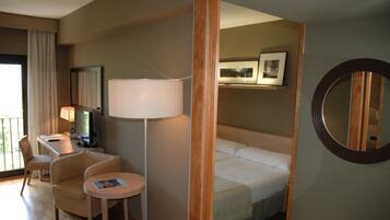 Habitación de hotel con cama, escritorio con computadora, silla, mesita, lámpara y espejo redondo.