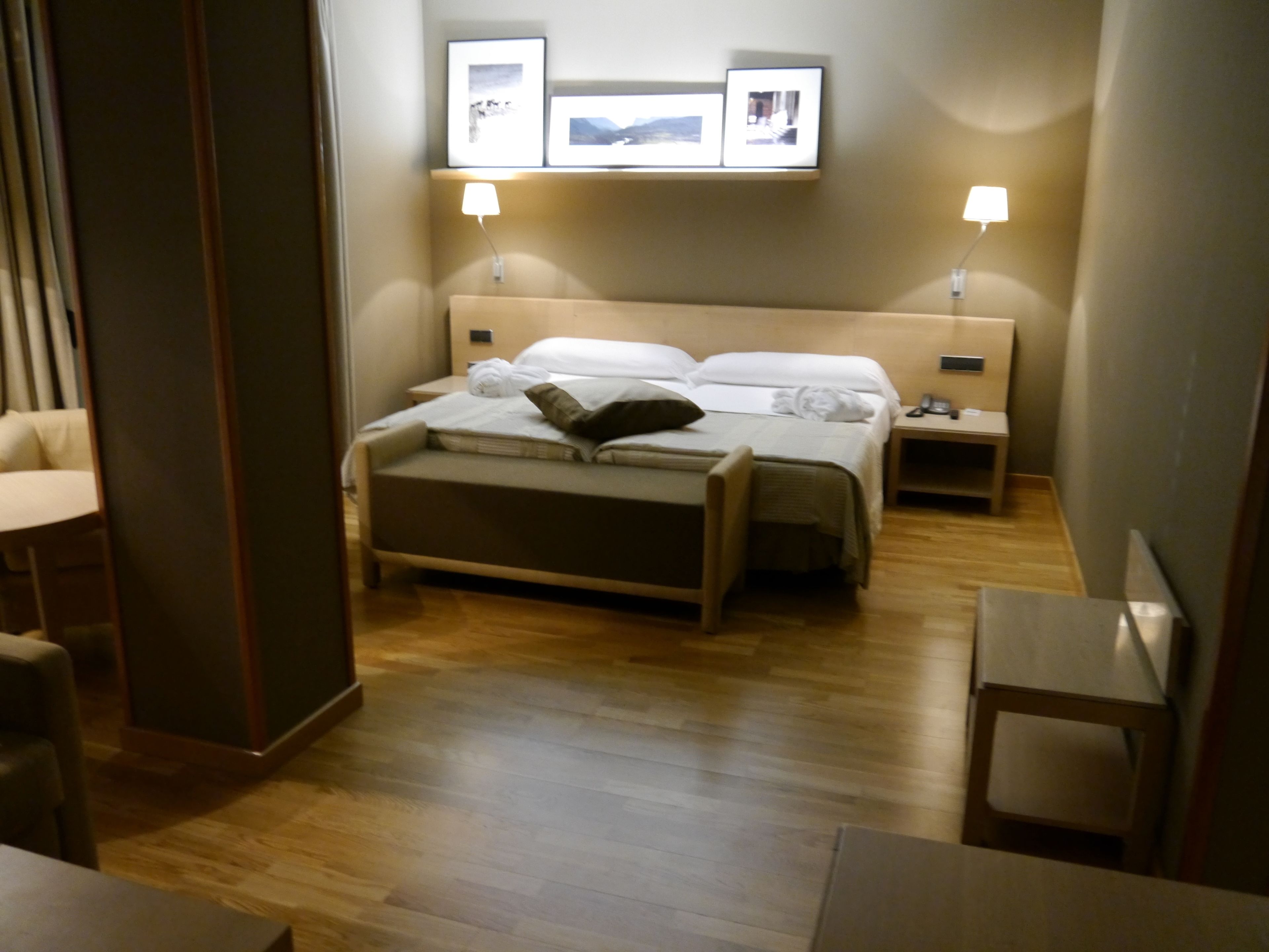 A hotel room with a bed, bedside tables, a desk, and a chair. There are framed pictures on the wall above the bed.
