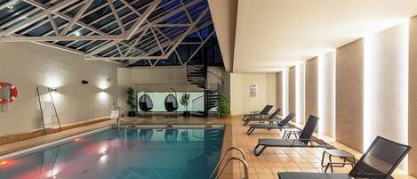 Una piscina interior con techo de cristal, tumbonas y una escalera.
