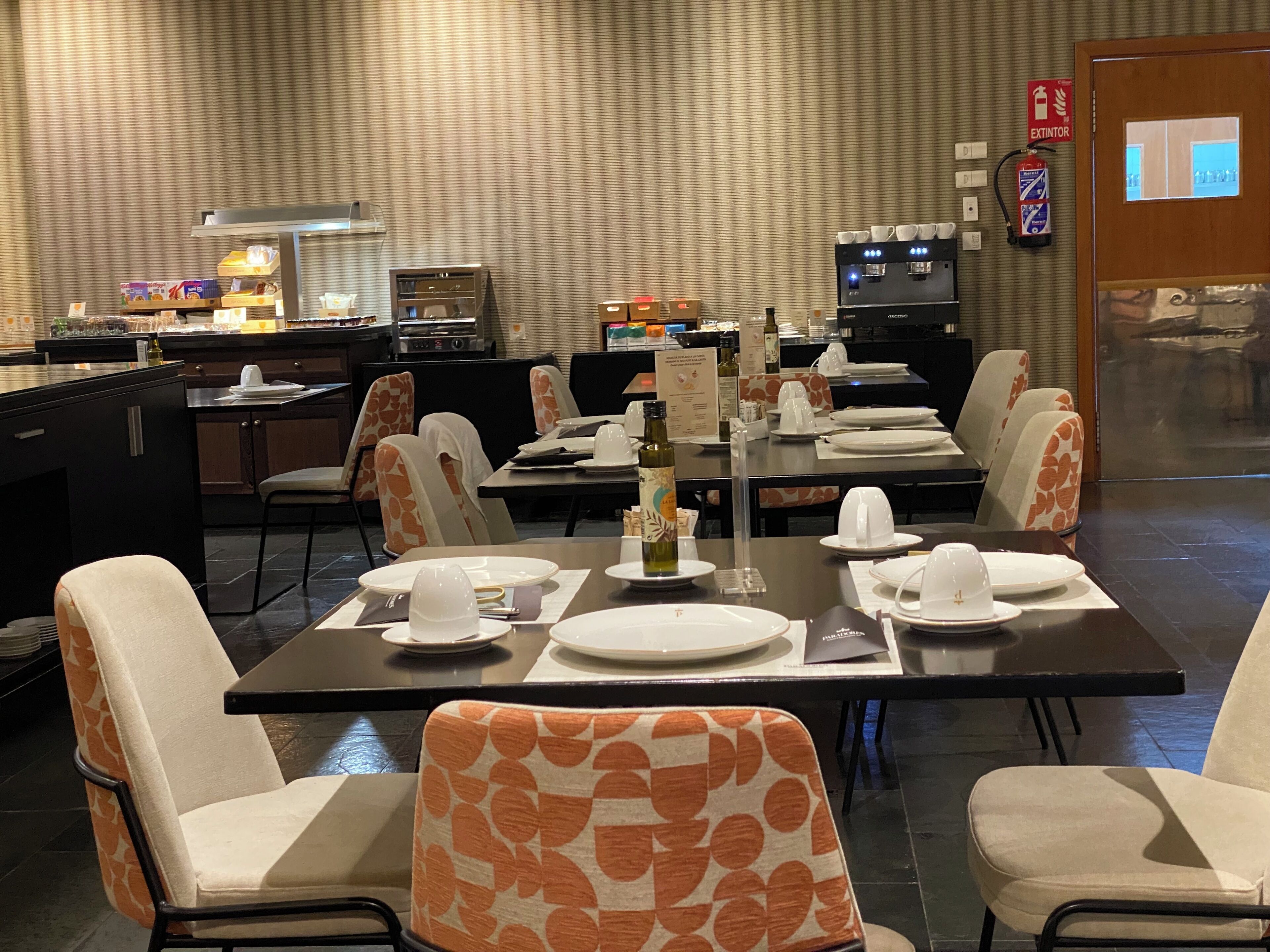 A restaurant with tables set for dining, chairs with orange patterns, a coffee machine, and an extinguisher sign on the wall.