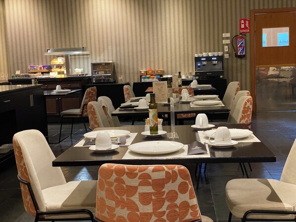A restaurant with tables set for dining, chairs with orange patterns, a coffee machine, and an extinguisher sign on the wall.