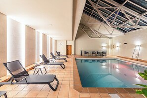 An indoor swimming pool with tiled flooring, black lounge chairs, and a high ceiling with skylights.