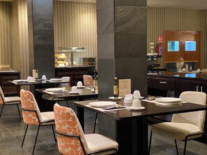 A modern dining area with high tables and chairs, a kitchen in the background, and a fire extinguisher on the wall.
