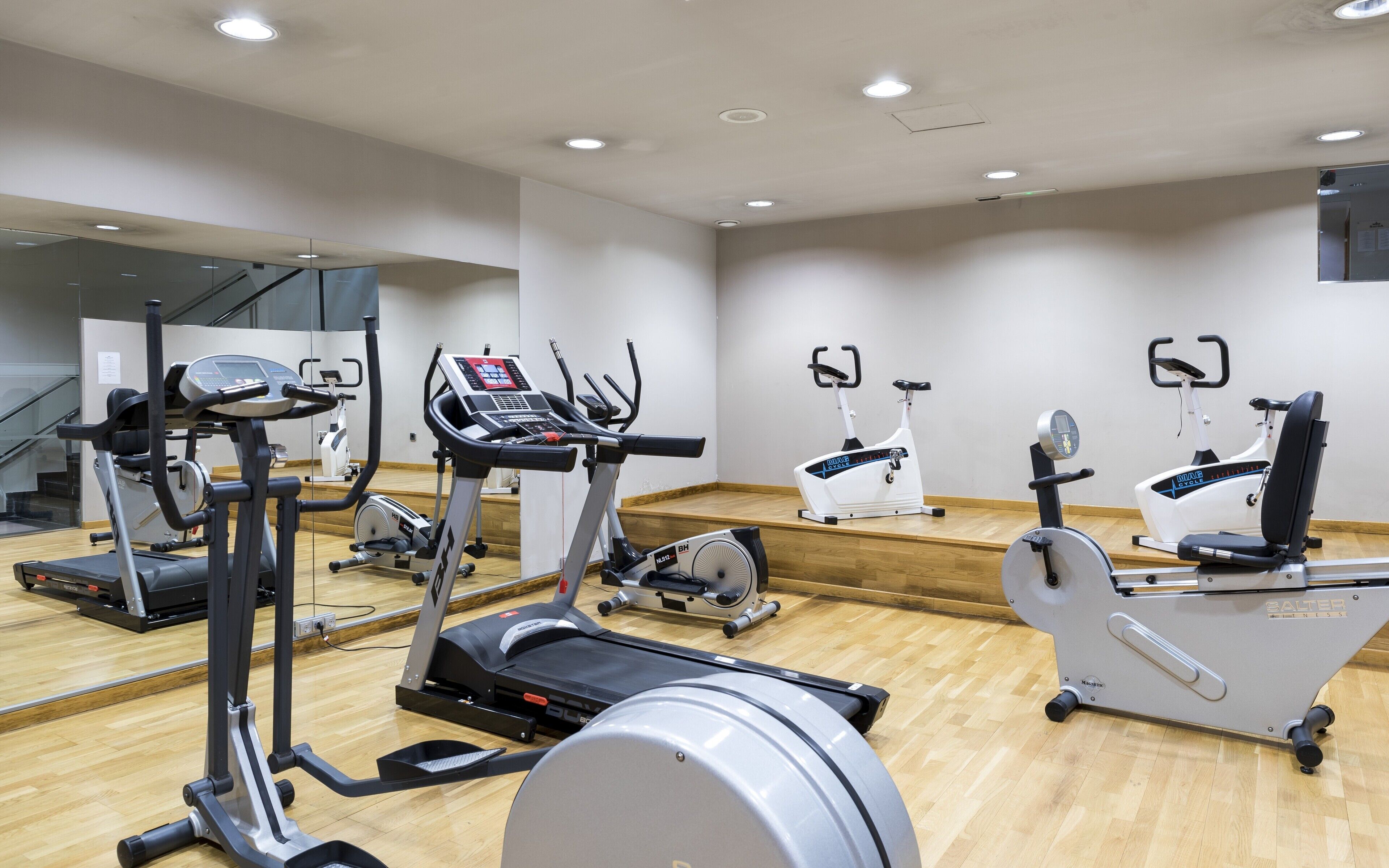 Un gimnasio con diferentes máquinas de cardio, piso de madera y un espejo grande.